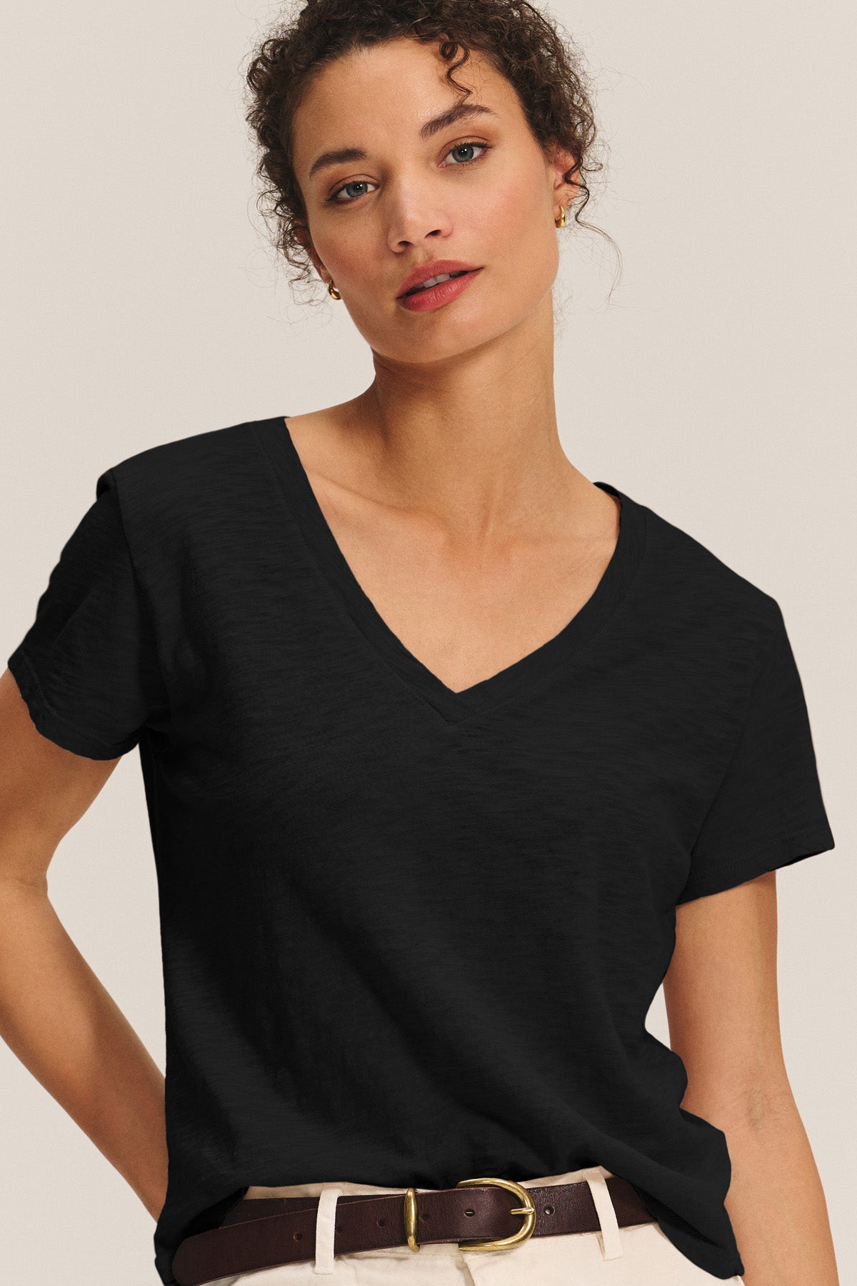 A woman wears the ELISHA TEE by Velvet by Graham & Spencer—a black cotton slub V-neck with short sleeves—paired with cream pants and a brown belt, standing against a light background: a true wardrobe staple.