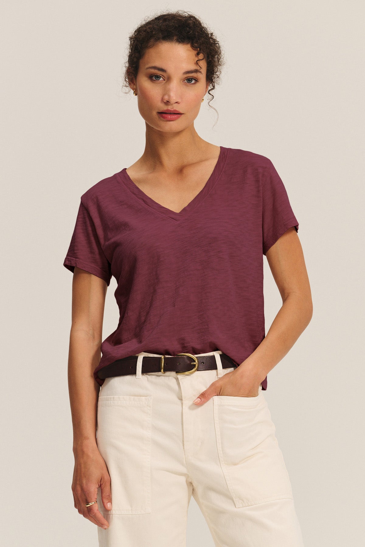 A woman wears the ELISHA TEE by Velvet by Graham & Spencer, a maroon cotton slub V-neck, tucked into high-waisted cream pants with a black belt, standing against a plain background.-40946868682945
