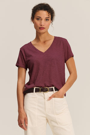 A woman wears the ELISHA TEE by Velvet by Graham & Spencer, a maroon cotton slub V-neck, tucked into high-waisted cream pants with a black belt, standing against a plain background.