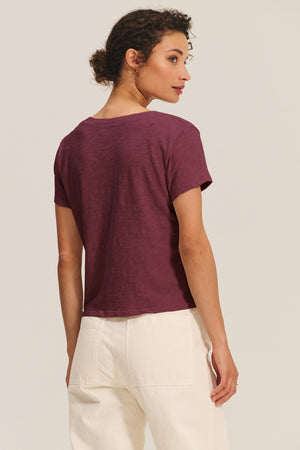 A person with curly hair is seen from the back wearing the Velvet by Graham & Spencer ELISHA TEE, a maroon cotton slub V-neck top, paired with high-waisted white pants against a plain background.