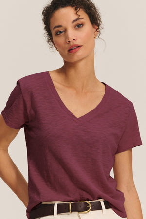 A woman in the ELISHA TEE by Velvet by Graham & Spencer—a maroon cotton slub V-neck—paired with beige pants and a brown belt stands against a plain background.