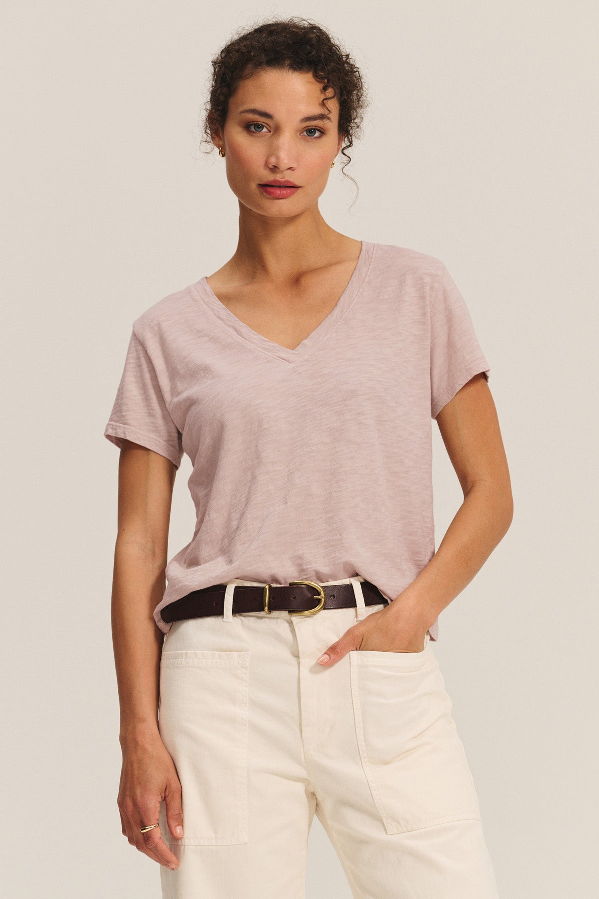 A woman faces forward wearing the ELISHA TEE by Velvet by Graham & Spencer—a light pink, soft cotton slub V-neck—tucked into high-waisted cream pants with a black belt, set against a plain background.