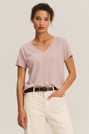 A woman faces forward wearing the ELISHA TEE by Velvet by Graham & Spencer—a light pink, soft cotton slub V-neck—tucked into high-waisted cream pants with a black belt, set against a plain background.