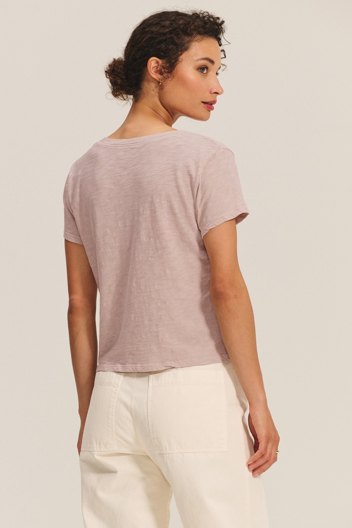A woman faces away, wearing the Velvet by Graham & Spencer ELISHA TEE in light mauve cotton slub with high-waisted white pants, set against a neutral background.