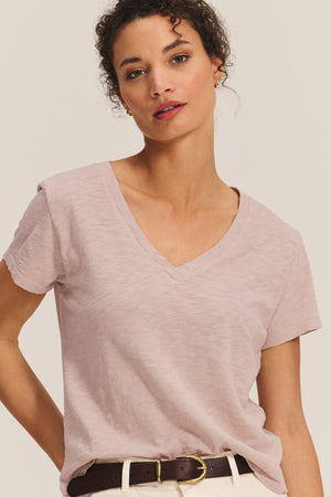 A person wears the ELISHA TEE by Velvet by Graham & Spencer, a light pink cotton slub V-neck, paired with white pants and a dark brown belt against a plain background.