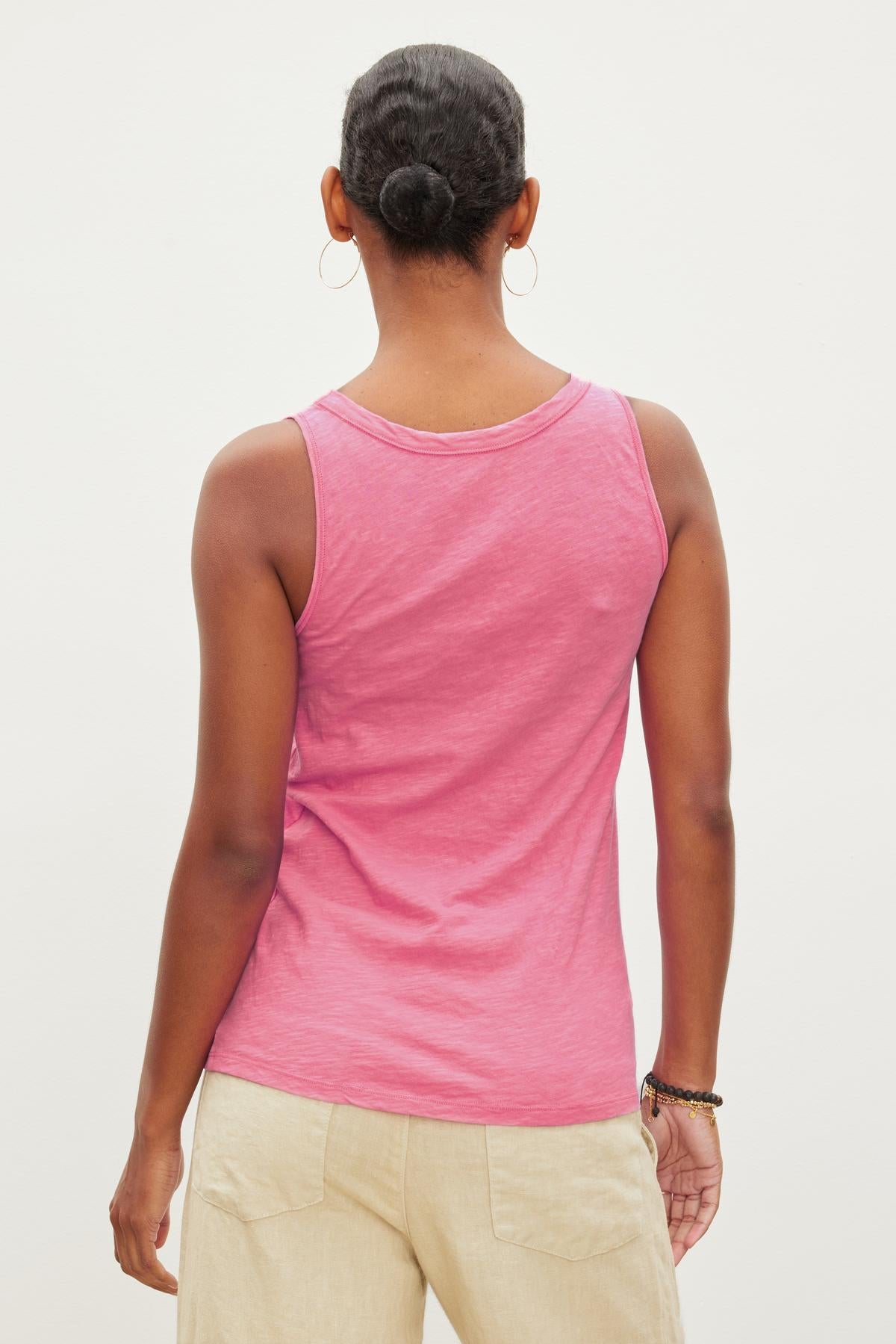   A person with tied-back hair is shown from the back, wearing a pink sleeveless JOY ORIGINAL SLUB SCOOP NECK TANK by Velvet by Graham & Spencer with flattering arm holes and beige pants, standing against a plain light background. 