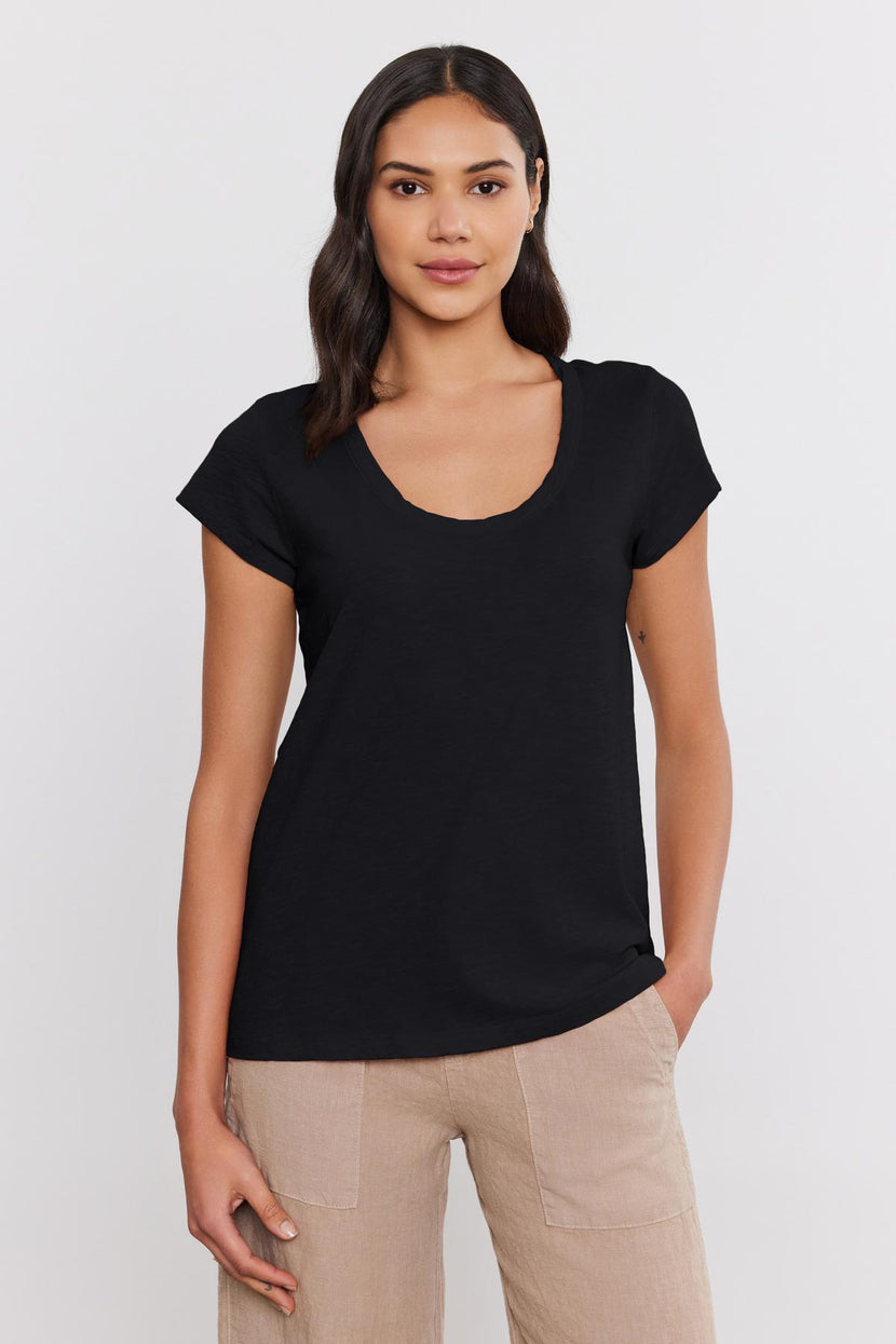 A woman with long dark hair is wearing a black *KIRA TEE* by *Velvet by Graham & Spencer* featuring a scoop neckline and beige pants, standing against a plain white background. Her outfit embodies everyday wear made comfortable with its cotton slub fabric, making it perfect for any casual occasion.