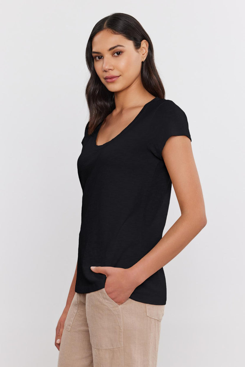 A person with long dark hair wears a black KIRA TEE by Velvet by Graham & Spencer made of cotton slub fabric and beige pants, standing against a plain white background.