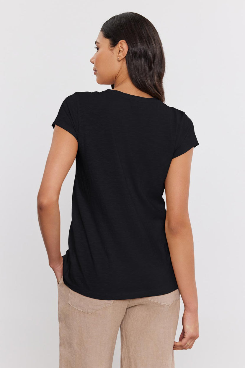 A woman with dark hair stands with her back to the camera, wearing a short-sleeve black KIRA TEE made of cotton slub fabric from Velvet by Graham & Spencer and beige pants, perfect for everyday wear.