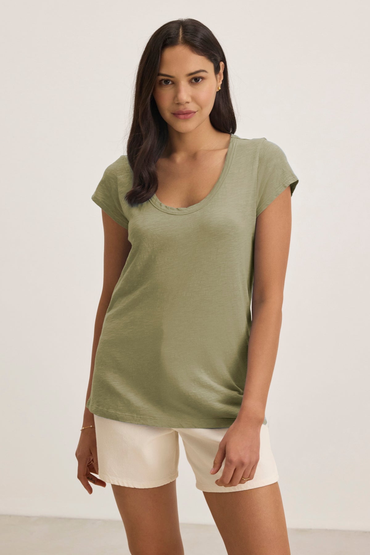 A woman stands against a plain background wearing the KIRA TEE by Velvet by Graham & Spencer, a light green cotton slub fabric t-shirt with cap sleeves, paired with off-white shorts.-39149282492609