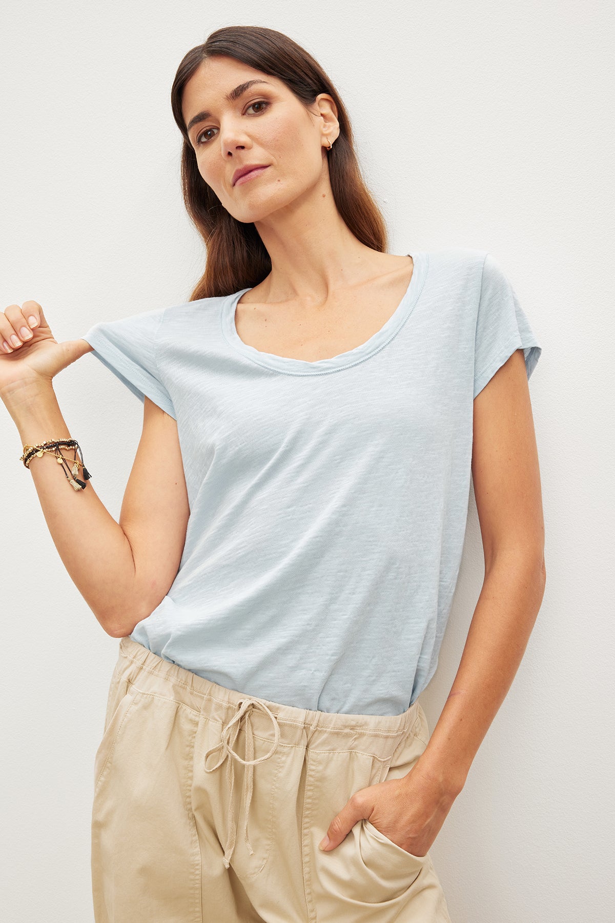 Woman in a light blue Velvet by Graham & Spencer KIRA ORIGINAL SLUB SCOOP NECK TEE with vintage-cropped sleeves and beige pants standing against a white wall, looking at the camera.-35982850490561