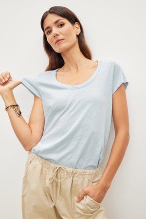 Woman in a light blue Velvet by Graham & Spencer KIRA ORIGINAL SLUB SCOOP NECK TEE with vintage-cropped sleeves and beige pants standing against a white wall, looking at the camera.