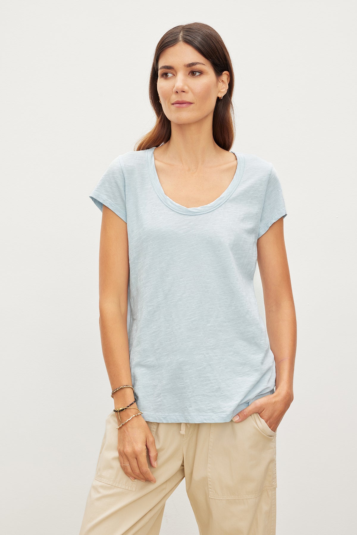   Woman in a light blue KIRA ORIGINAL SLUB SCOOP NECK TEE by Velvet by Graham & Spencer with vintage-cropped sleeves and scoop neck, paired with beige pants, standing against a plain background, looking slightly to the side. 