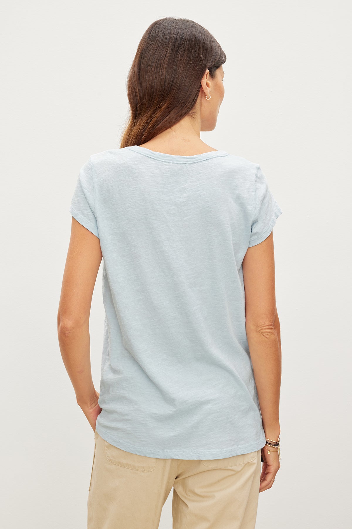   Woman viewed from behind, wearing a light blue Velvet by Graham & Spencer KIRA ORIGINAL SLUB SCOOP NECK TEE and beige pants, standing against a plain white background. 