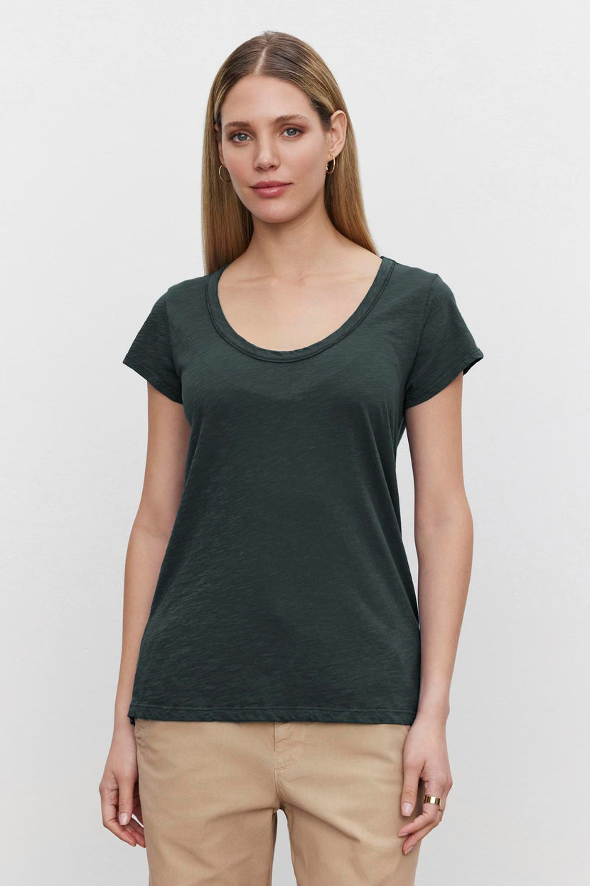 An individual with long hair is seen standing against a plain white background, wearing the KIRA TEE—a dark green top crafted from cotton slub fabric by Velvet by Graham & Spencer—paired with beige pants.