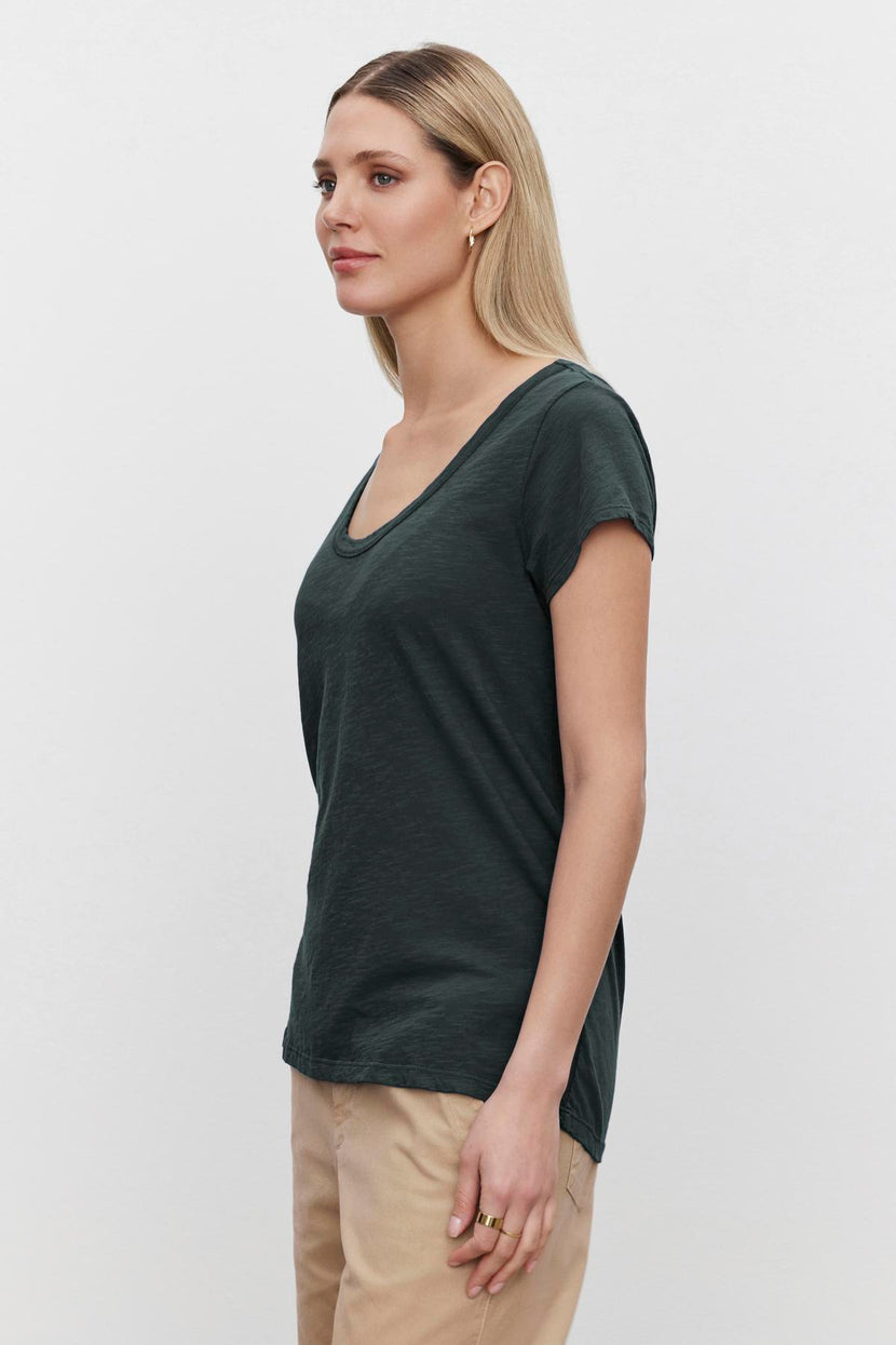 A woman with long hair stands in side profile, wearing the KIRA TEE by Velvet by Graham & Spencer, a dark green t-shirt featuring a scoop neckline and cap sleeves, paired with beige pants against a plain white background.