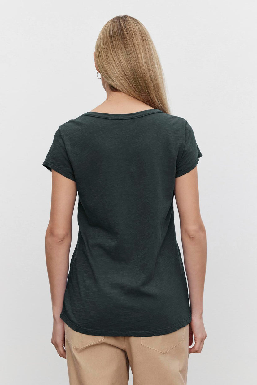 Rear view of a person with long blonde hair wearing the KIRA TEE, a dark green cap sleeve T-shirt by Velvet by Graham & Spencer, paired with beige pants against a white background.