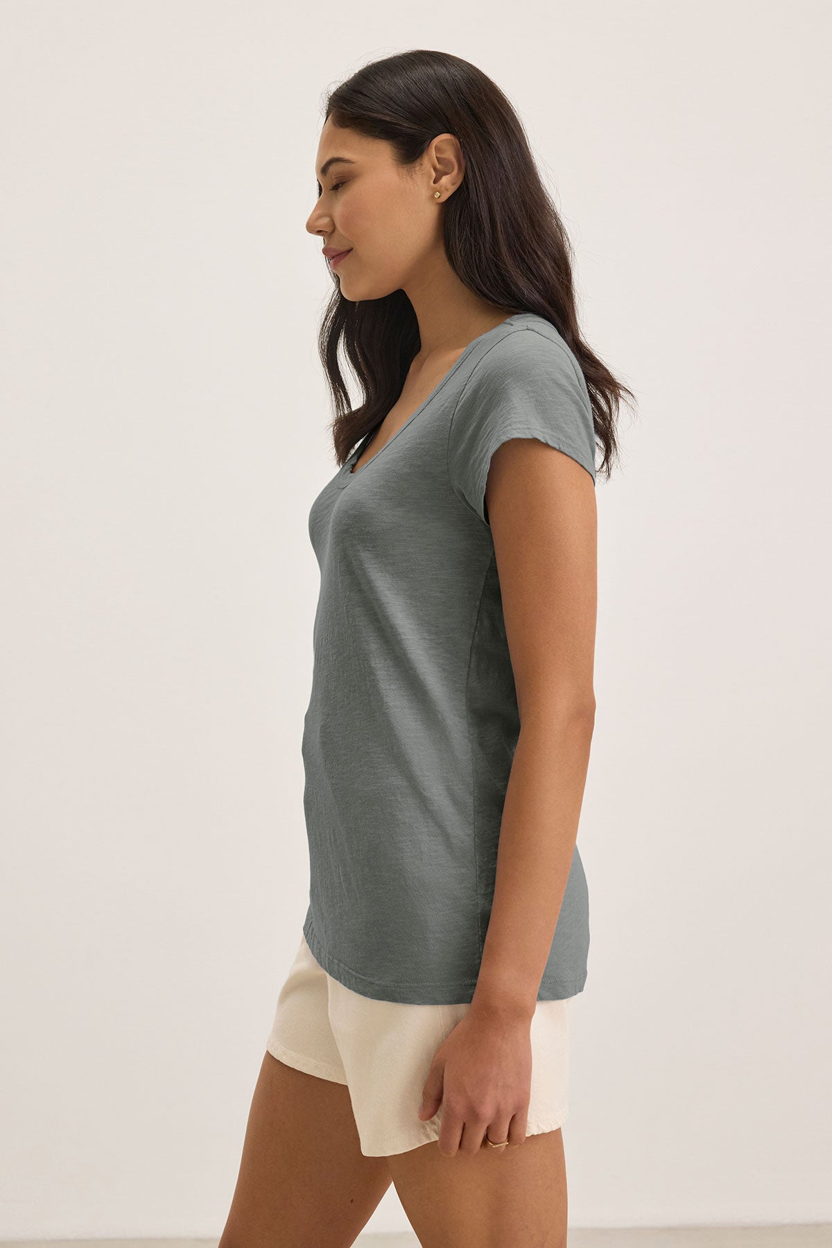   A woman in a Velvet by Graham & Spencer KIRA TEE, featuring a green scoop neckline, and beige shorts stands in profile against a plain background, showcasing the effortless style of everyday wear. 