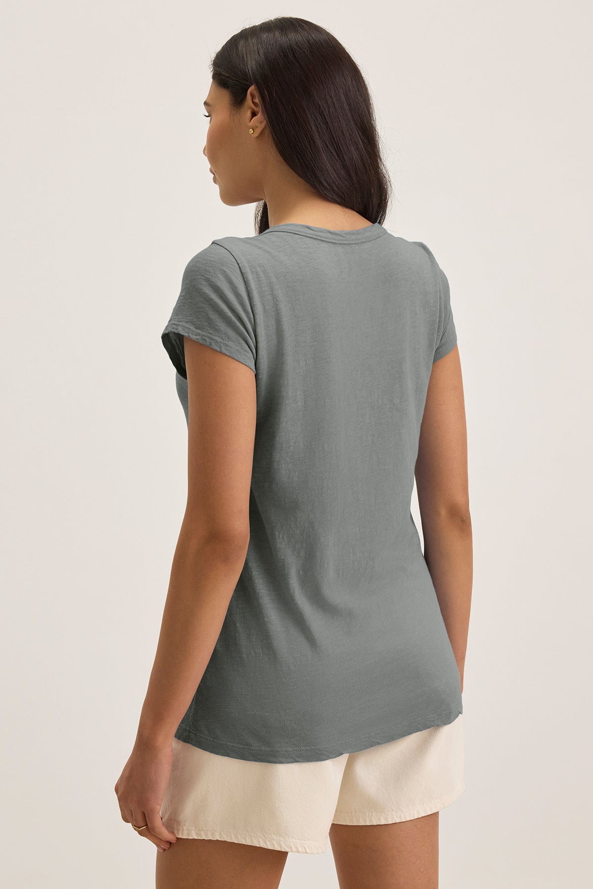   A person with long dark hair dons the KIRA TEE, a green scoop neck T-shirt by Velvet by Graham & Spencer, paired with beige shorts made from soft cotton slub fabric. They gaze into the distance, perfectly blending comfort and style for everyday wear. 