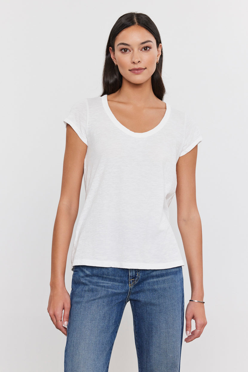 A person with long dark hair, wearing a white short-sleeve V-neck KIRA TEE from Velvet by Graham & Spencer made from a soft cotton slub fabric and blue jeans, stands against a plain white background.