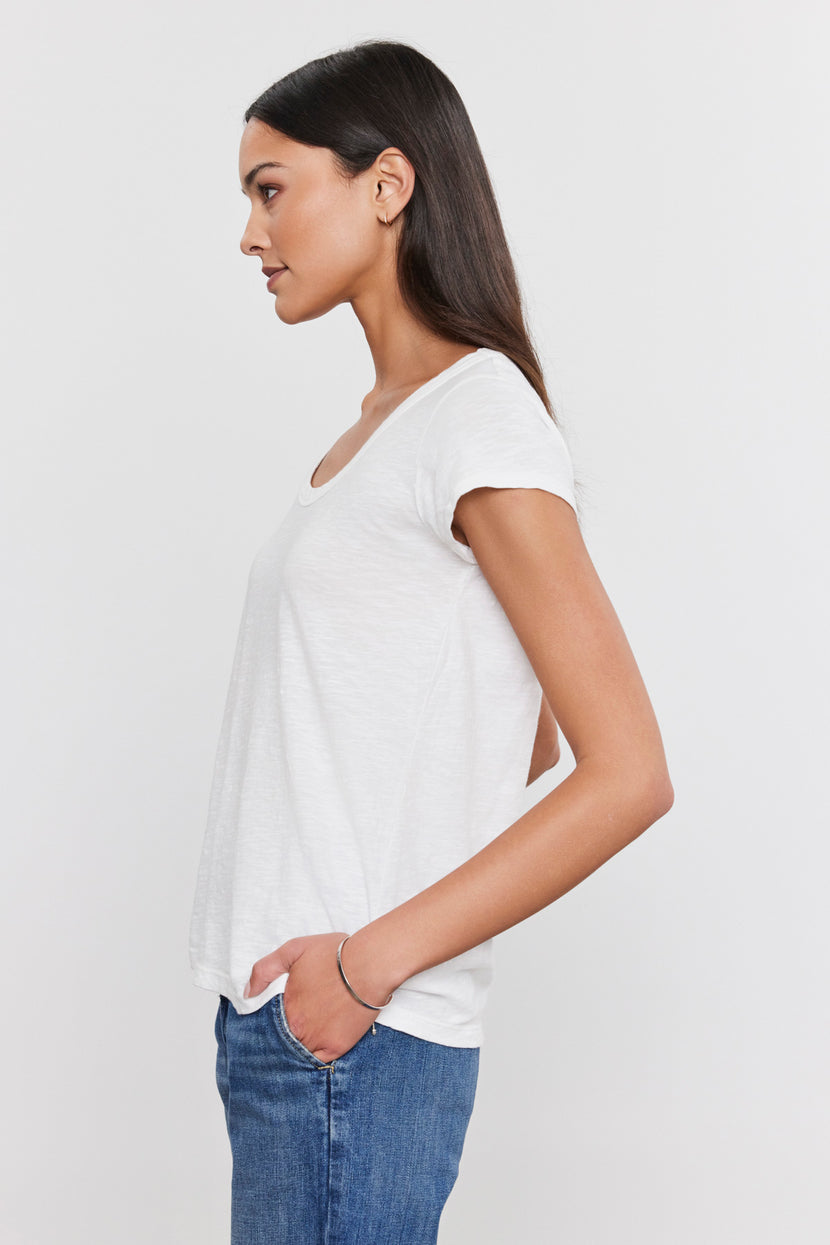 Side profile of a person with long, dark hair wearing a white t-shirt with a scoop neckline and blue jeans, looking forward and placing their left hand in their pocket against a plain white background. Perfected everyday wear crafted from soft cotton slub fabric: the KIRA TEE by Velvet by Graham & Spencer.