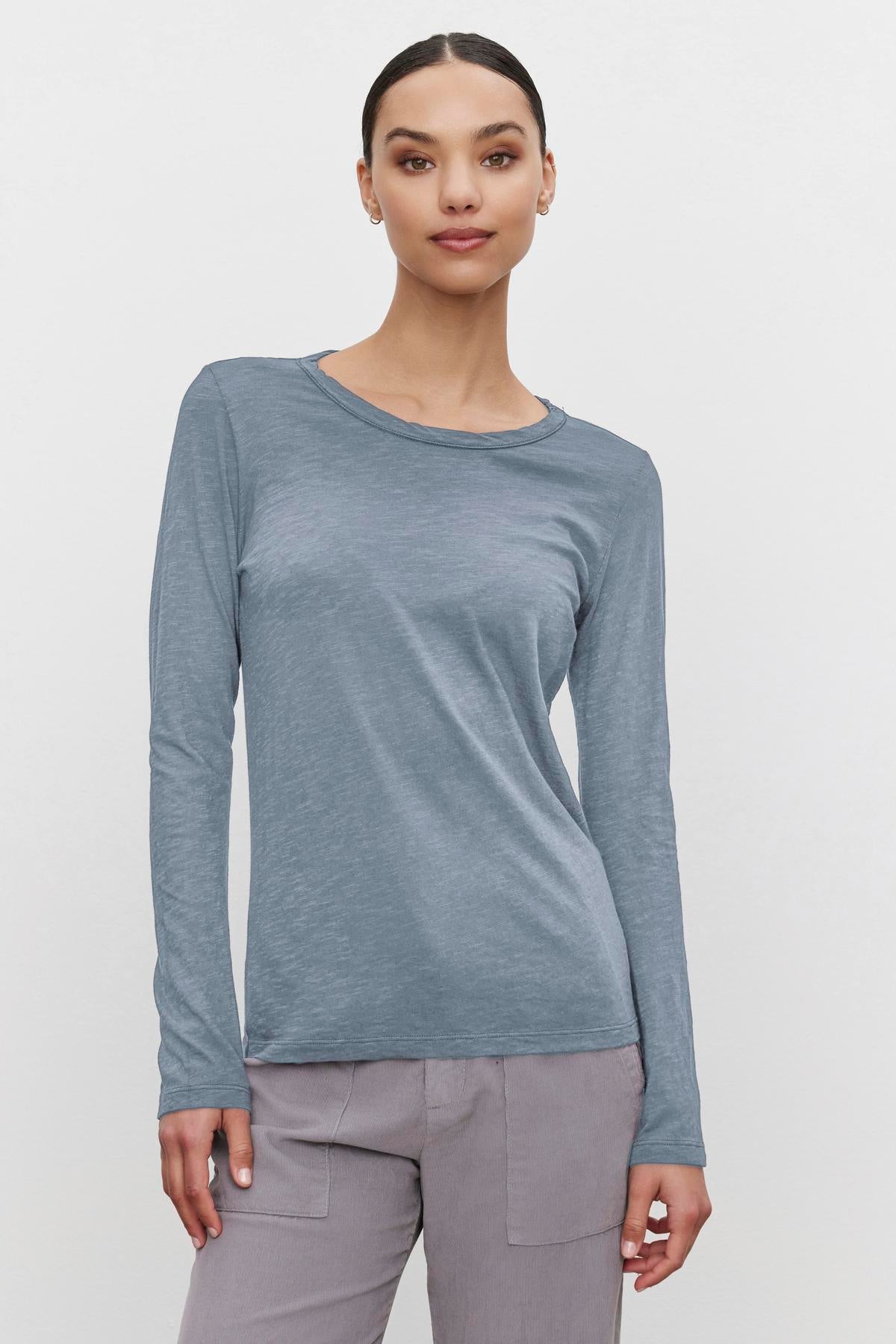   A person wearing the LIZZIE TEE, a blue long-sleeve shirt by Velvet by Graham & Spencer, with a classic crew neckline and gray pants, standing against a plain white background. 