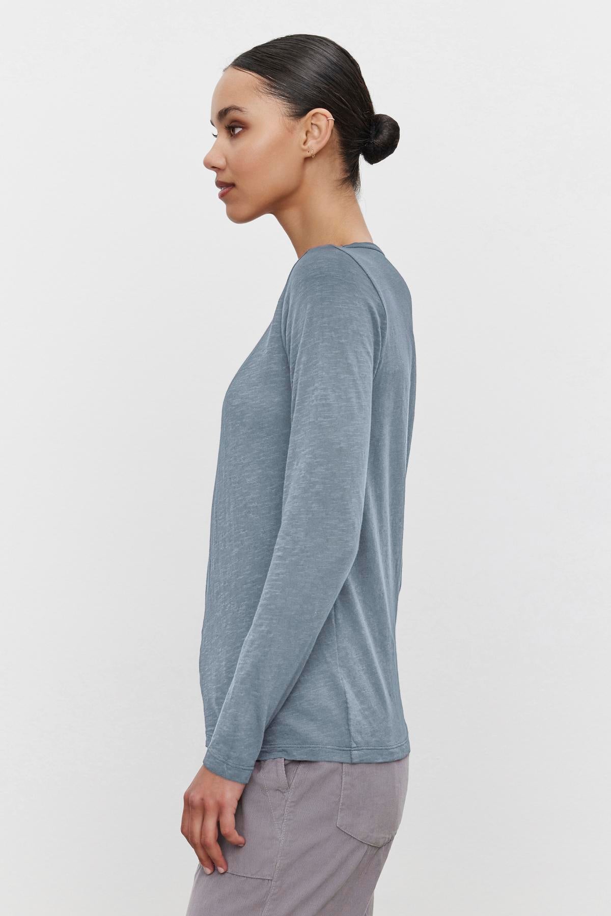   A person in profile wearing the LIZZIE TEE, a gray long-sleeve tee by Velvet by Graham & Spencer, with a classic crew neckline and gray pants, standing against a white background. 