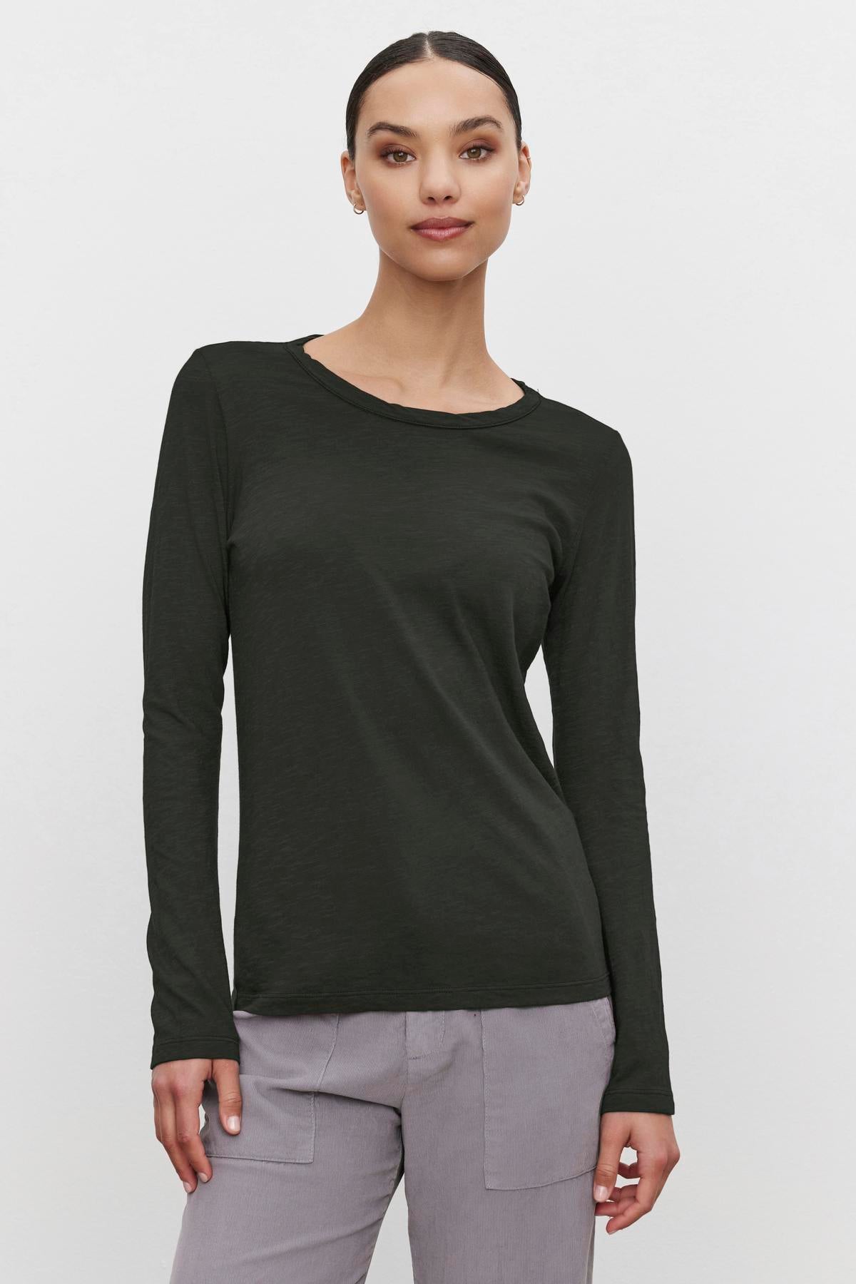   A person wearing the LIZZIE TEE by Velvet by Graham & Spencer, a long-sleeved dark green shirt with a classic crew neckline, paired with gray pants, stands against a plain white background. 