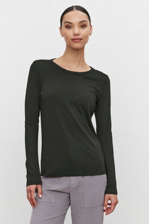 A person wearing the LIZZIE TEE by Velvet by Graham & Spencer, a long-sleeved dark green shirt with a classic crew neckline, paired with gray pants, stands against a plain white background.