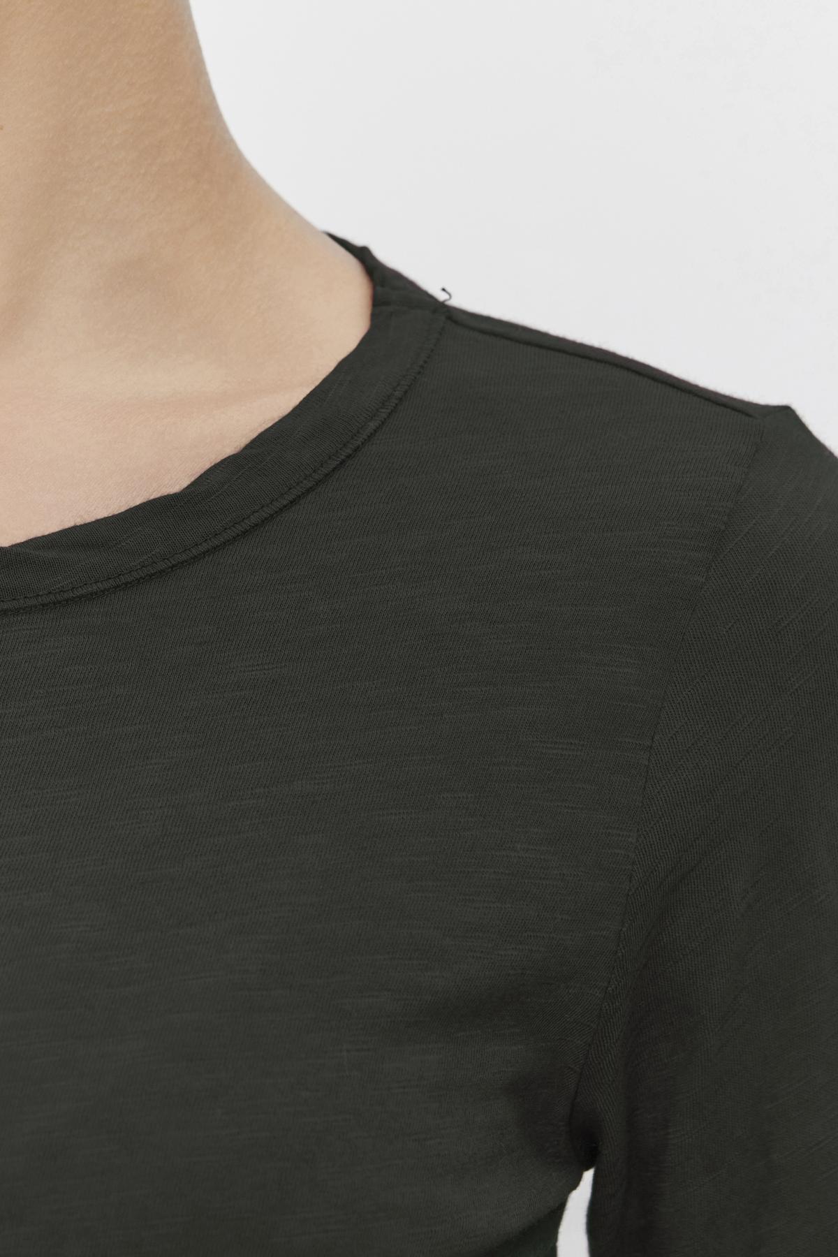 Close-up of a person wearing the LIZZIE TEE by Velvet by Graham & Spencer, featuring a classic crew neckline in dark green. The image highlights the shoulder and part of the upper chest against a neutral background, showcasing the depth and character added by its textured cotton slub fabric to this simple yet stylish piece.-38333534142657