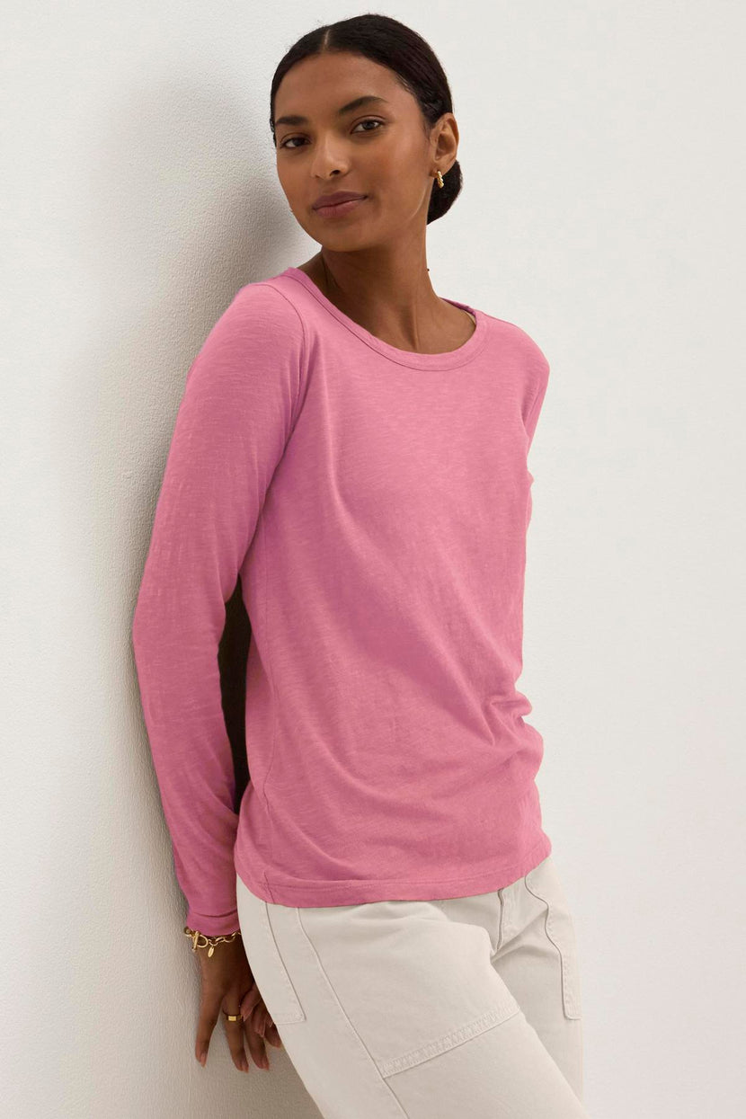 A person wearing the Velvet by Graham & Spencer LIZZIE TEE in pink cotton slub, paired with white pants, leans against a white wall while looking at the camera.