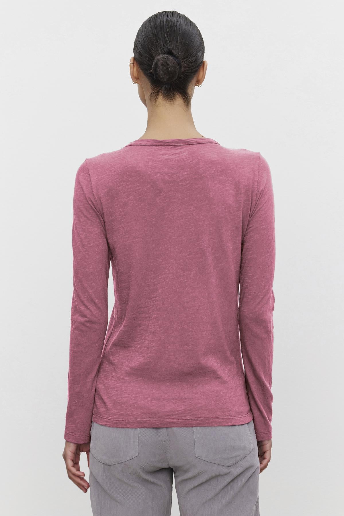 A person with a bun wears the Velvet by Graham & Spencer LIZZIE TEE, a long-sleeved pink shirt with a classic crew neckline, paired with gray pants, shown from the back against a plain white background.-38642774671553