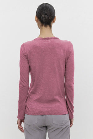 A person with a bun wears the Velvet by Graham & Spencer LIZZIE TEE, a long-sleeved pink shirt with a classic crew neckline, paired with gray pants, shown from the back against a plain white background.