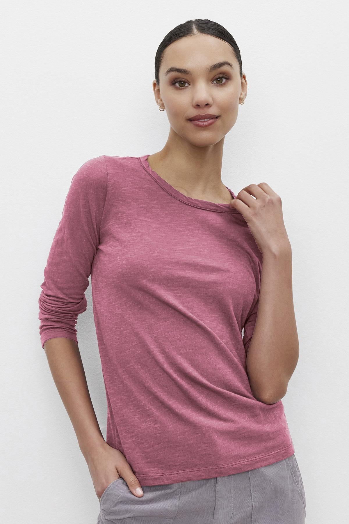   Someone wearing the Velvet by Graham & Spencer LIZZIE TEE, a long-sleeve pink top with a classic crew neckline, and gray pants stands against a plain white background, holding the neckline with one hand and resting the other in a pocket. 