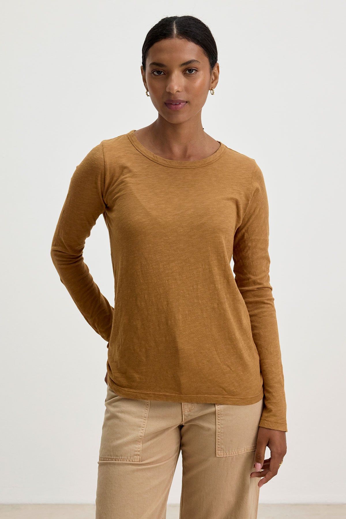 A person with dark hair is wearing the long-sleeve, mustard-colored "LIZZIE TEE" by Velvet by Graham & Spencer, along with beige pants, and is standing against a plain background.-37745662689473