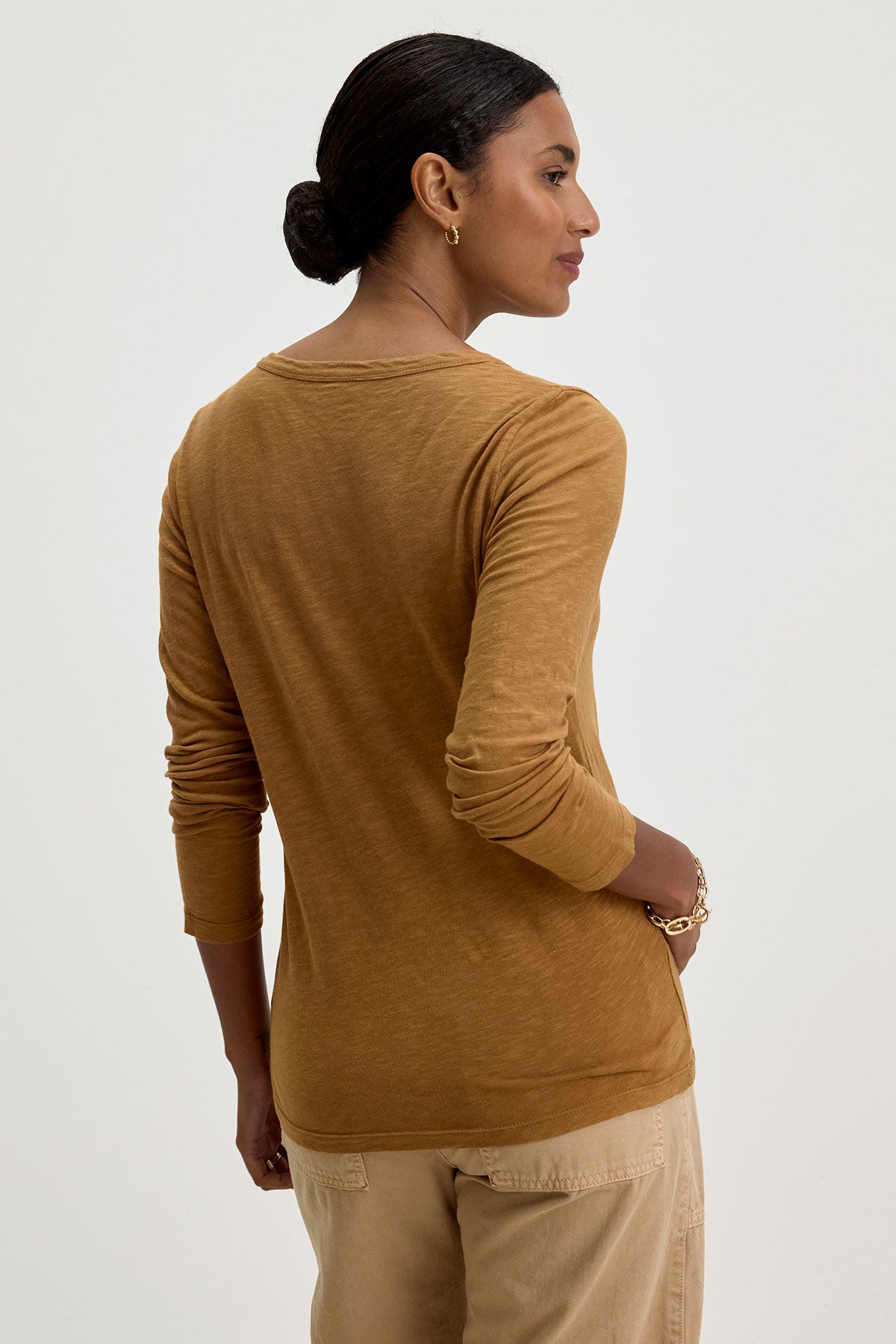   A person with dark hair tied back wears the LIZZIE TEE from Velvet by Graham & Spencer, a long-sleeve, mustard-colored top featuring a classic crew neckline. They have one hand on their hip and are facing away from the camera. 