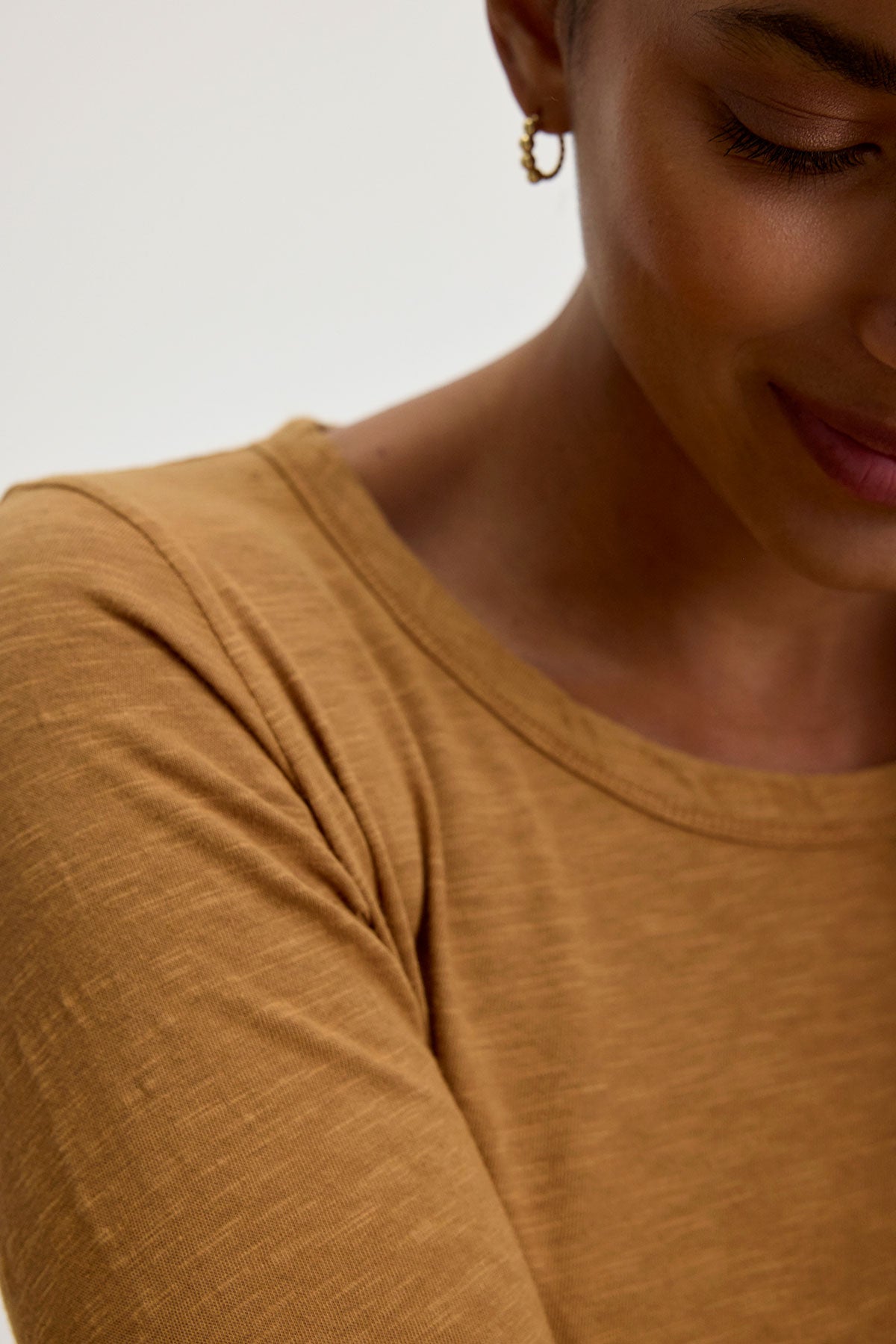   A person wearing the LIZZIE TEE by Velvet by Graham & Spencer—featuring a mustard color, classic crew neckline, and long sleeves—smiles gently, showcasing one small hoop earring against a plain background. 