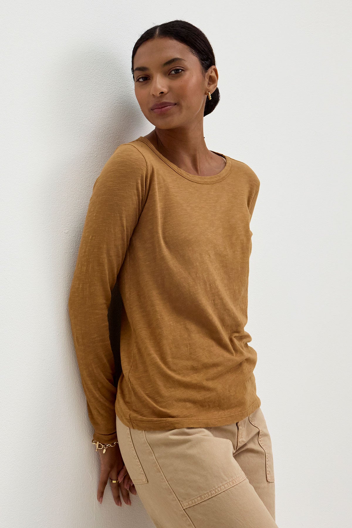 A person with short dark hair, wearing the LIZZIE TEE from Velvet by Graham & Spencer, a long-sleeved brown top with a classic crew neckline, paired with beige pants, leans against a white wall.-37745662656705