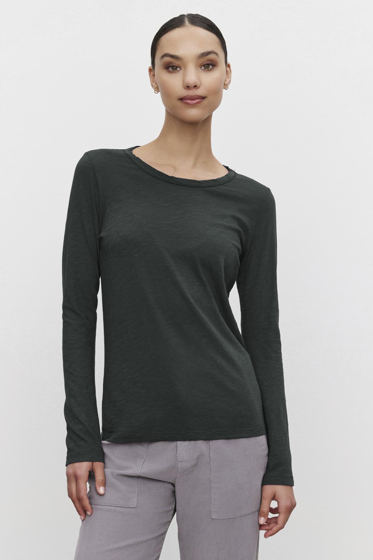   A person wearing a LIZZIE TEE by Velvet by Graham & Spencer, styled in dark green with a crew neckline and long sleeves, along with gray pants, stands against a plain background. 