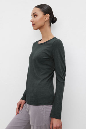 Someone wearing the LIZZIE TEE, a dark green long-sleeve shirt with a crew neckline from Velvet by Graham & Spencer, stands against a plain white background.