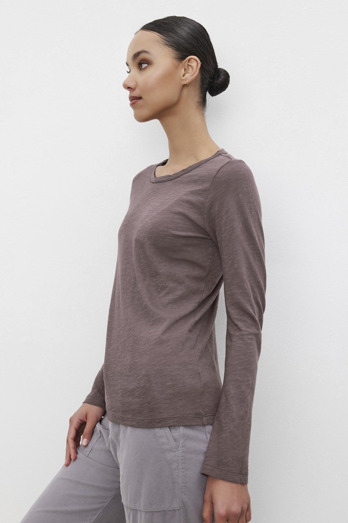 A person with hair styled in a bun stands against a white background, wearing the Long-Sleeve LIZZIE TEE by Velvet by Graham & Spencer, featuring a classic crew neckline, paired with gray pants.-37935126053057