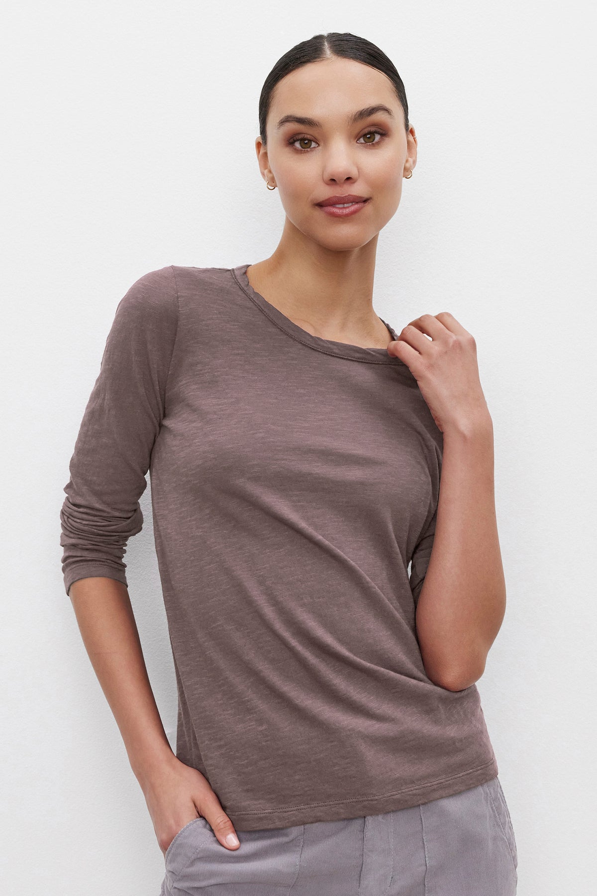   Person wearing the LIZZIE TEE, a long-sleeve, brown shirt by Velvet by Graham & Spencer, featuring a classic crew neckline. One hand touches the collar while the other rests in a pocket as they stand against a plain white background. 