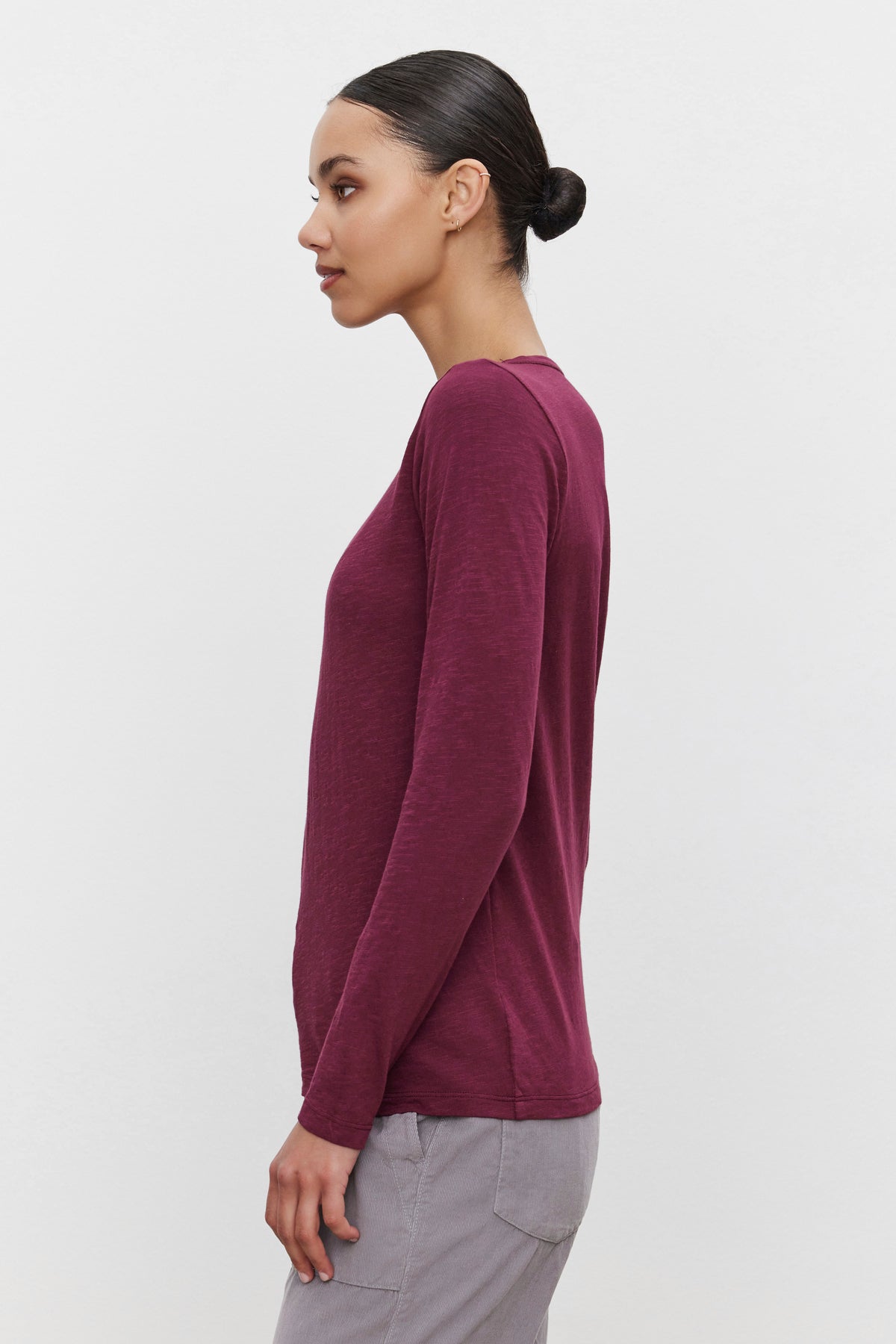   Side profile of a person with tied-back hair, wearing the maroon LIZZIE TEE by Velvet by Graham & Spencer, made of textured cotton slub, and light gray pants, standing against a plain white background. 