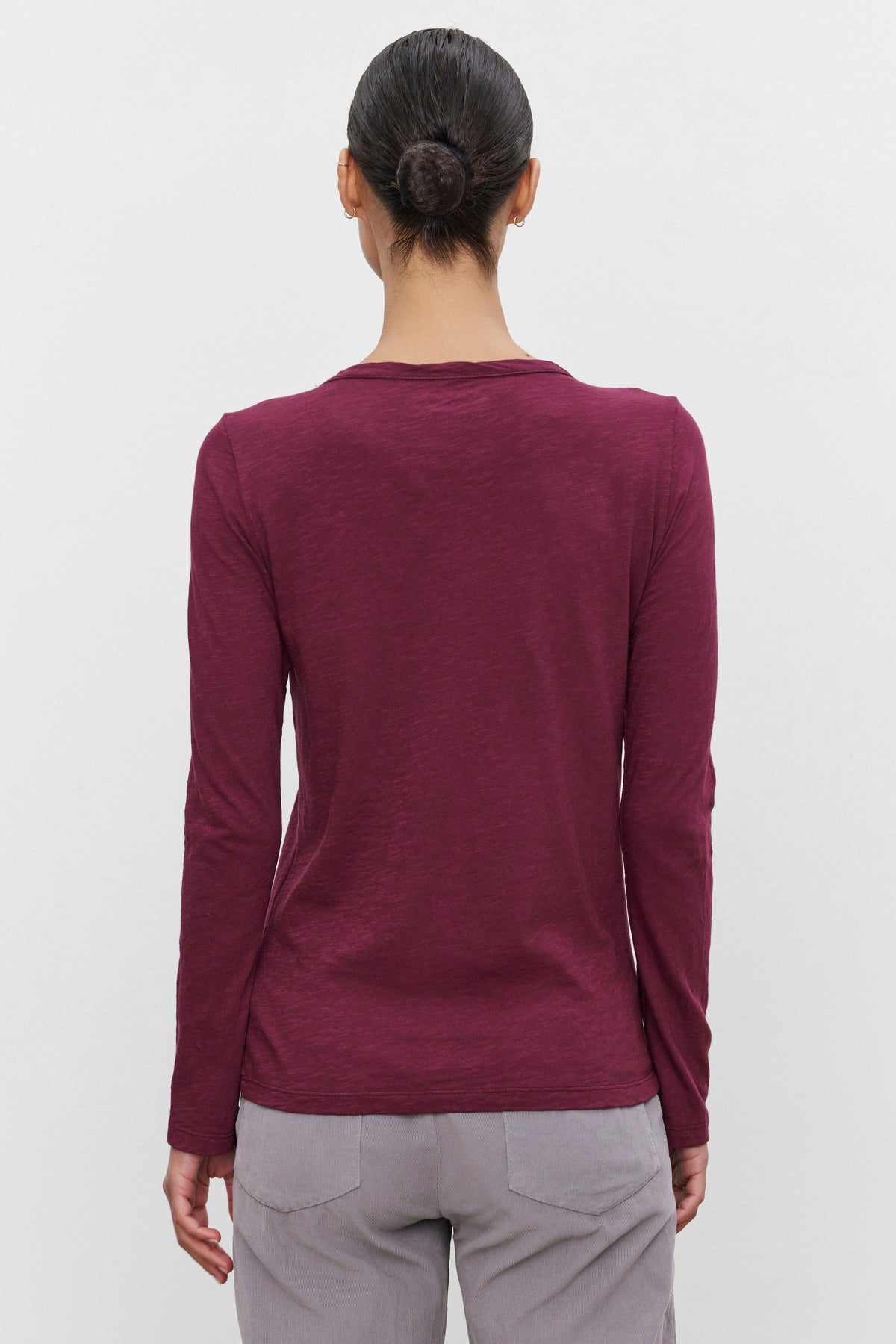   A person with dark hair in a bun is shown from the back, wearing the LIZZIE TEE by Velvet by Graham & Spencer — a maroon shirt with a classic crew neckline and long sleeves, paired with light gray pants. 