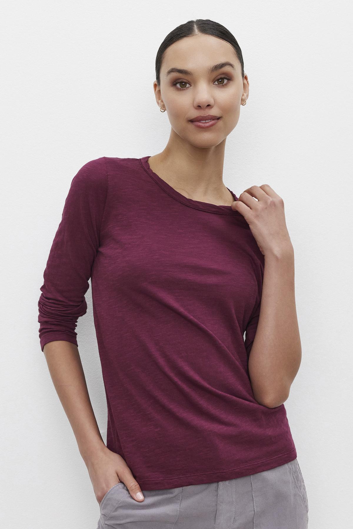 A person with dark hair tied back, wearing the LIZZIE TEE by Velvet by Graham & Spencer—a long-sleeved, textured cotton slub maroon shirt—and light gray pants, poses against a plain white background with one hand in their pocket and the other touching their collar.-37676904808641