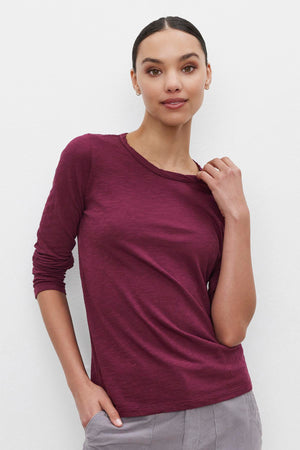 A person with dark hair tied back, wearing the LIZZIE TEE by Velvet by Graham & Spencer—a long-sleeved, textured cotton slub maroon shirt—and light gray pants, poses against a plain white background with one hand in their pocket and the other touching their collar.