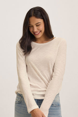 A person wearing the LIZZIE TEE by Velvet by Graham & Spencer, a cream long-sleeve top with a crew neckline, paired with blue jeans, is smiling and looking down against a plain background.