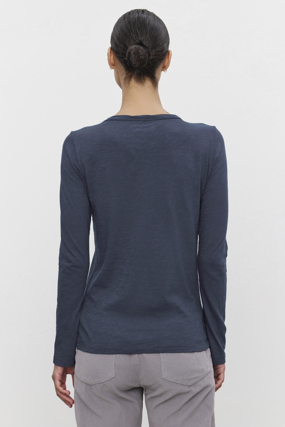   A person with dark hair in a bun is shown from the back, wearing the Velvet by Graham & Spencer LIZZIE TEE, a long-sleeve, dark blue shirt with a classic crew neckline, paired with light-colored pants. 