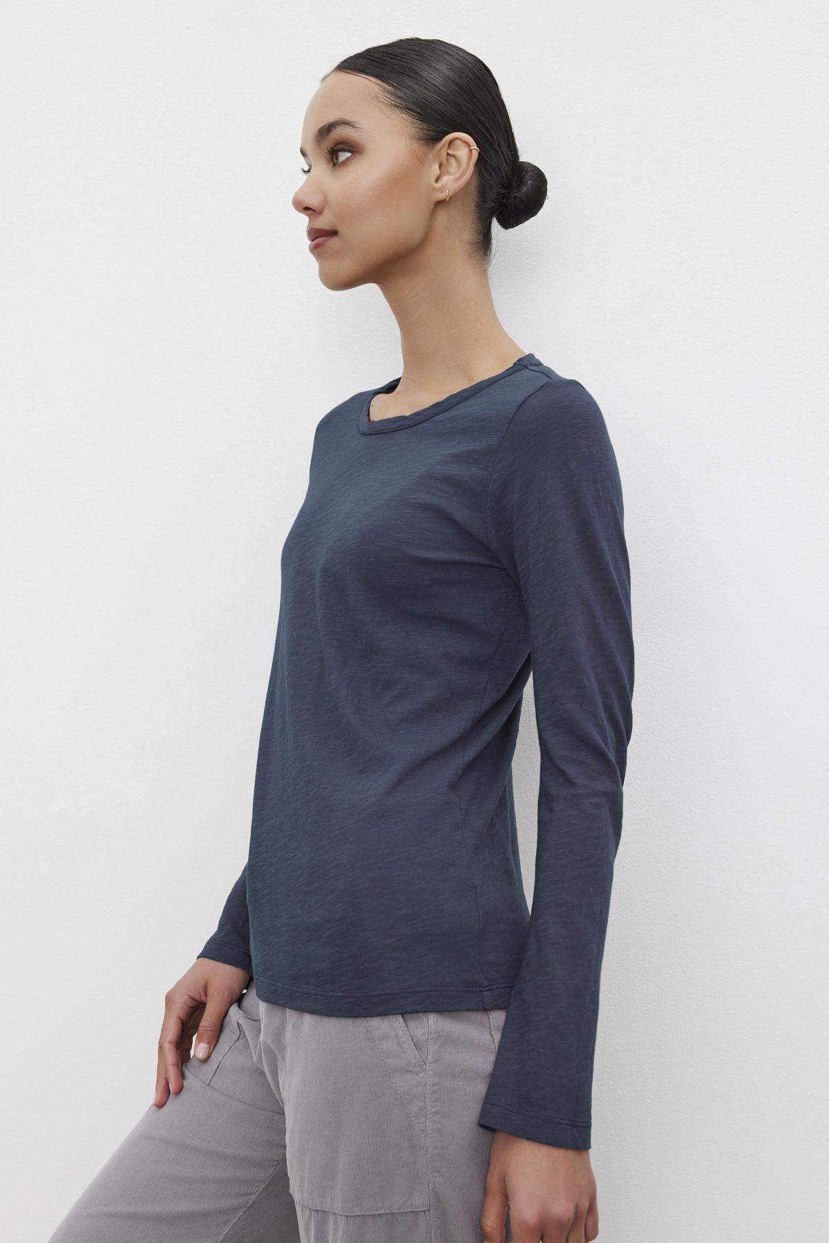   A person with their hair in a bun wears the Velvet by Graham & Spencer LIZZIE TEE, a long-sleeved, navy blue shirt with a classic crew neckline, paired with light-colored pants. They are turned slightly to the side against a plain white background. 