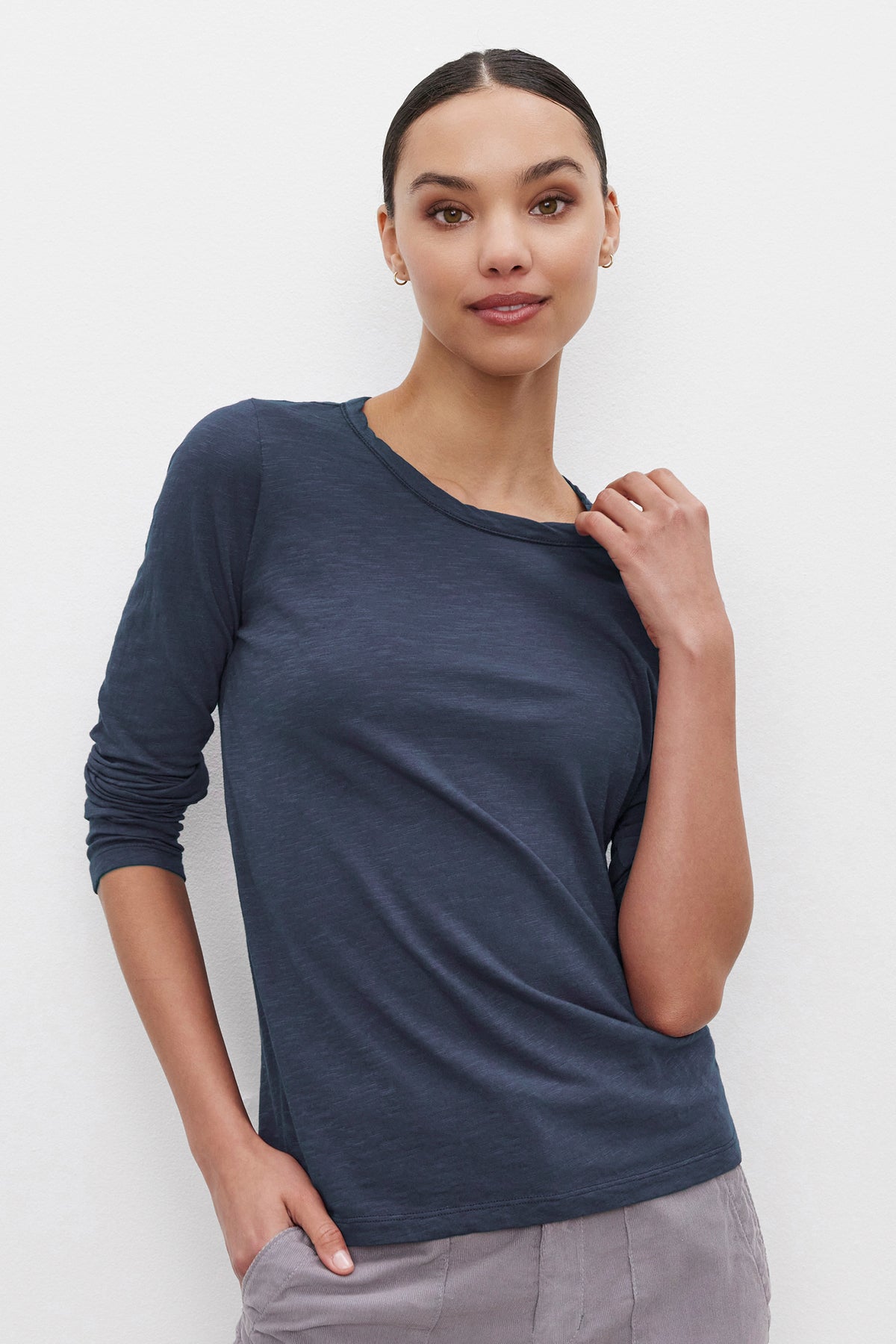 A person with dark, pulled-back hair, wearing the LIZZIE TEE from Velvet by Graham & Spencer – a classic crew neckline long-sleeve top in dark blue – paired with light-colored pants, stands against a light background, touching their collar with one hand and placing the other in their pocket.-37935125790913